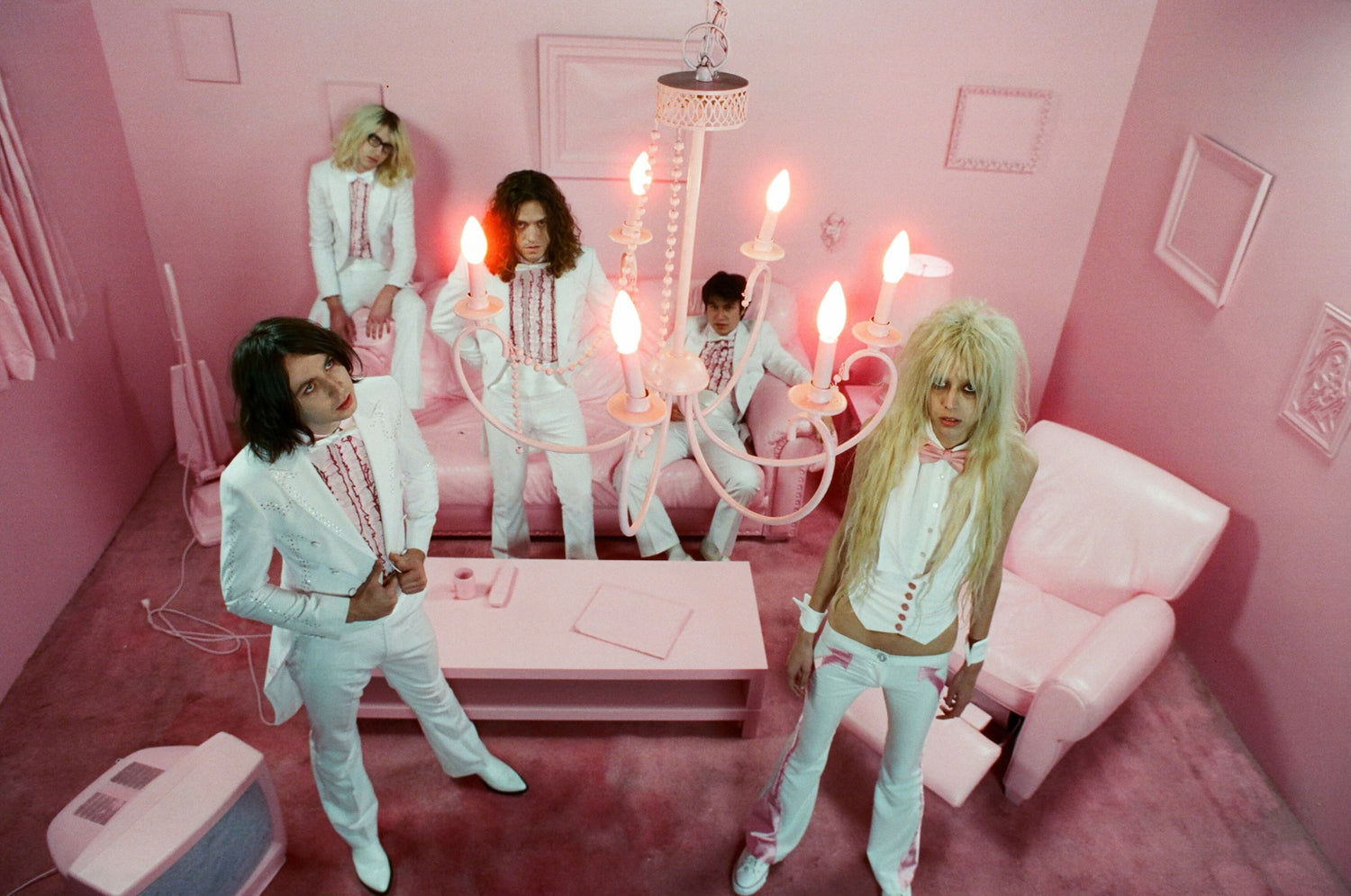 image of the five members of the band, starcrawler, all in white tuxedo outfits standing in a pink room with pink carpet, tv, chair, couch and under a chandelier looking up.