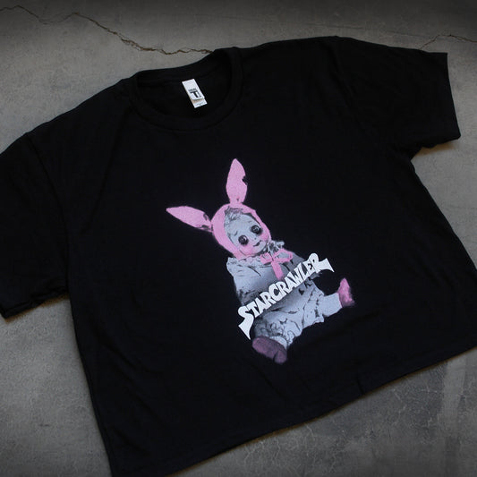 close up, angled image of a women's black cropped tee laid flat on a concrete floor. tee has center chest print of a baby doll sitting with pink bunny ears on its head. across the doll in white says starcrawler