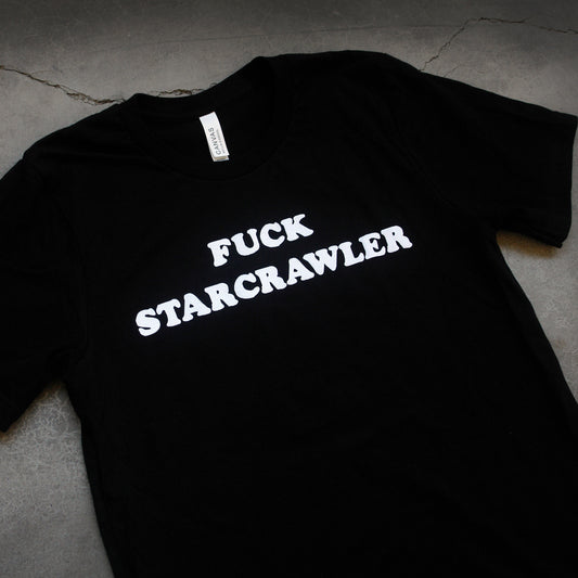 close up. angled image of a black tee laid flat on a concrete floor. white text across the chest that says fuck starcrawler