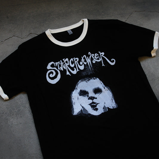 close up angled image of a black and white ringer tee laid flat on a concrete floor. front of tee has grey print of a spooky face. at the top across the chest says starcrawler