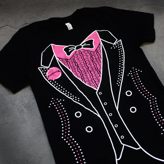 close up, angled image of a black tee laid flat on a concrete floor. tee has full body print to look like a tuxedo with a pink shirt, black tie, vest and opened jacket. small pink circle to represent a button on the left lapel that says starcrawler